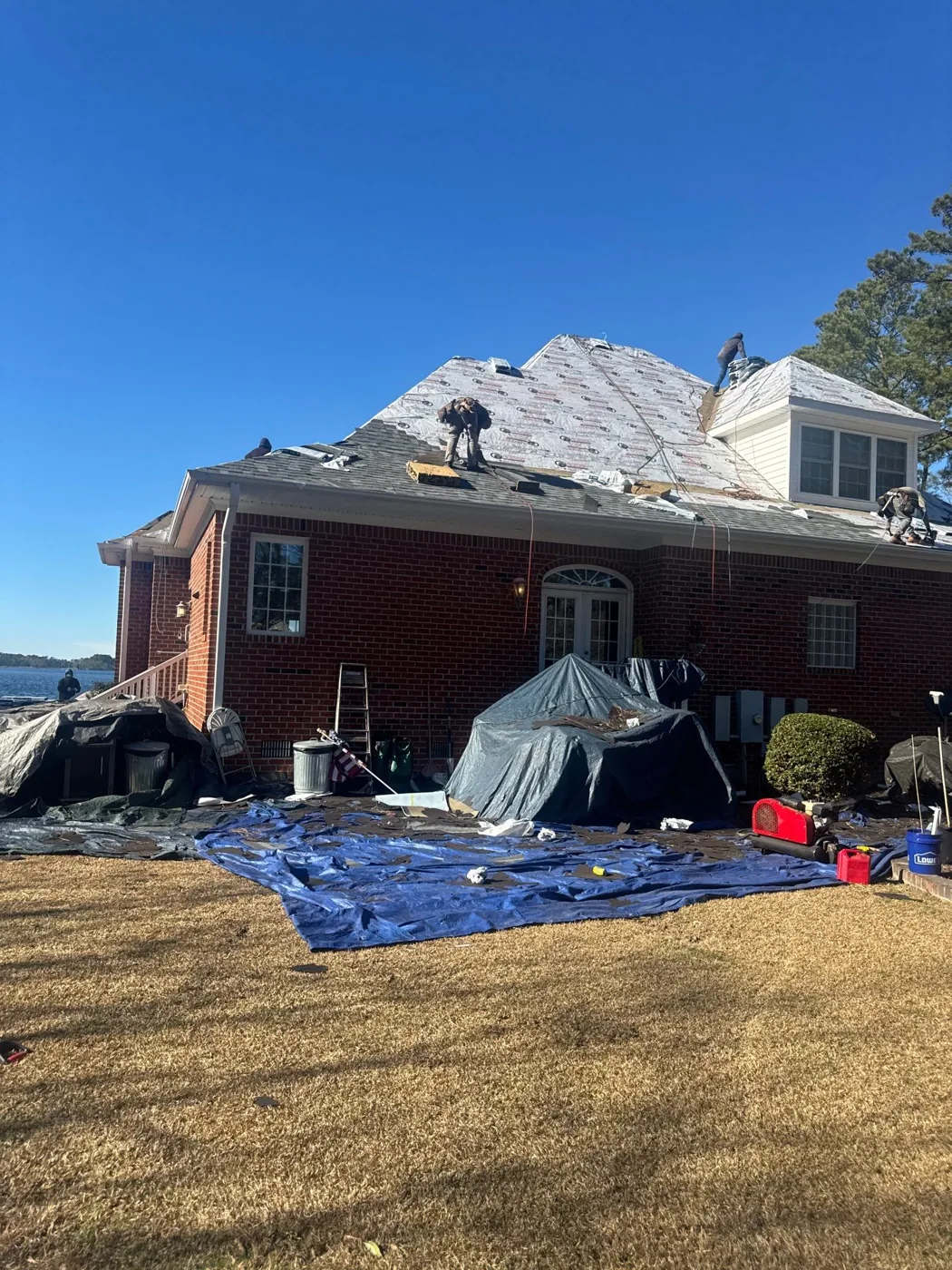 Red brick home mid re-roof, crew on the slope and grey tarps draped over the shrubs and beds below